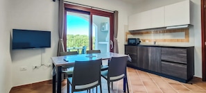 Een eethoek met een tafel en stoelen, een aan de muur bevestigde televisie, een keuken met een magnetron, en een glazen schuifdeur naar een balkon.