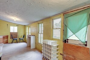 A room with wooden walls, a white ceiling, and a window with a green checkered blind. There are two chairs and a white shelf unit. - Home Near Eleven Point River & Nat'l Forest! (Alton)
