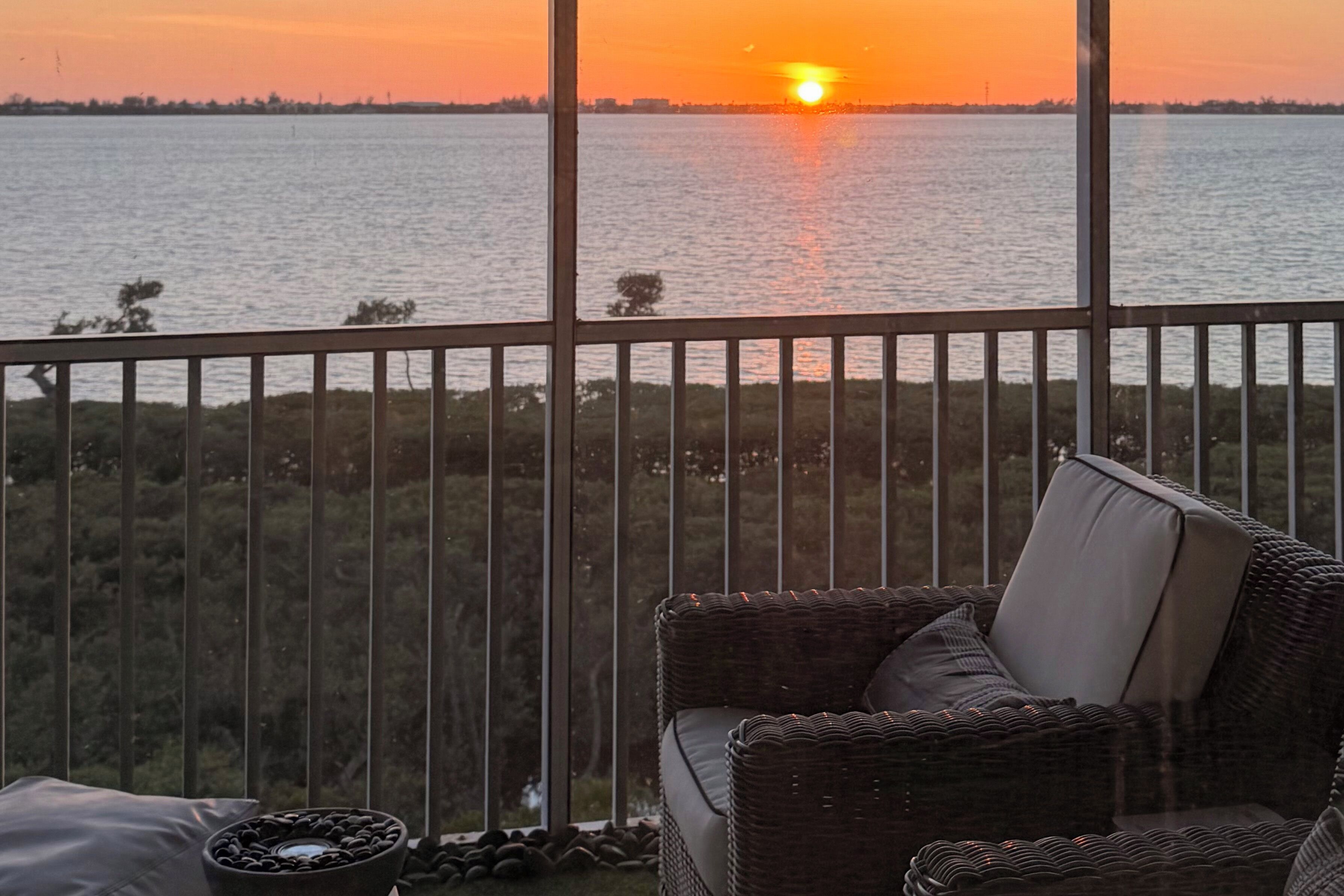 Een balkon met een rieten stoel en een klein tafeltje, uitkijkend op een wateroppervlak met een zonsondergang.