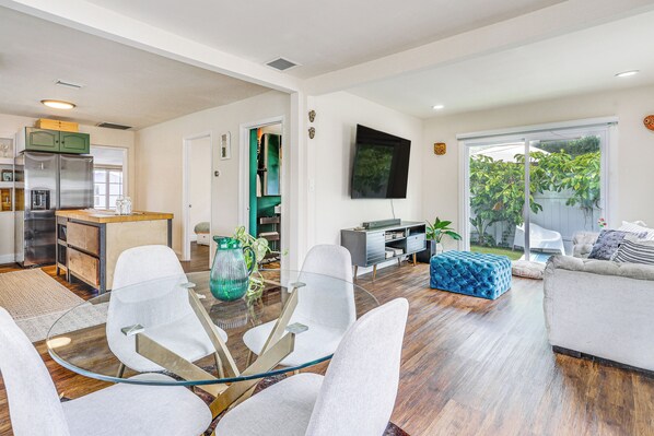 A modern living room with a glass coffee table, white chairs, a sofa, and a TV mounted on the wall.