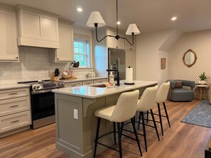 A modern kitchen with a central island, bar stools, a refrigerator, and a dining area with a sofa and side table.