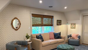 A living room with a beige sofa, a blue armchair, a green ottoman, a round side table with a plant, and a mirror.
