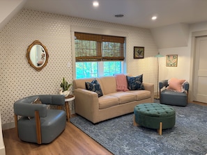 A living room with a beige sofa, a blue armchair, a green ottoman, a round side table with a plant, and a mirror.