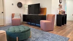 A living room with a television, two armchairs, a green ottoman, a patterned rug, and a black entertainment unit.