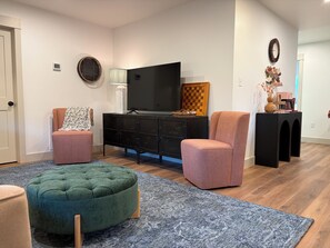 A living room with a television, two armchairs, a green ottoman, a patterned rug, and a black entertainment unit.