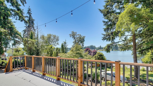 A deck with a wooden railing overlooking a lake, surrounded by trees and a clear sky. - Walk to Spanaway Lake: Chic Home w/ Rooftop Deck! (Spanaway)