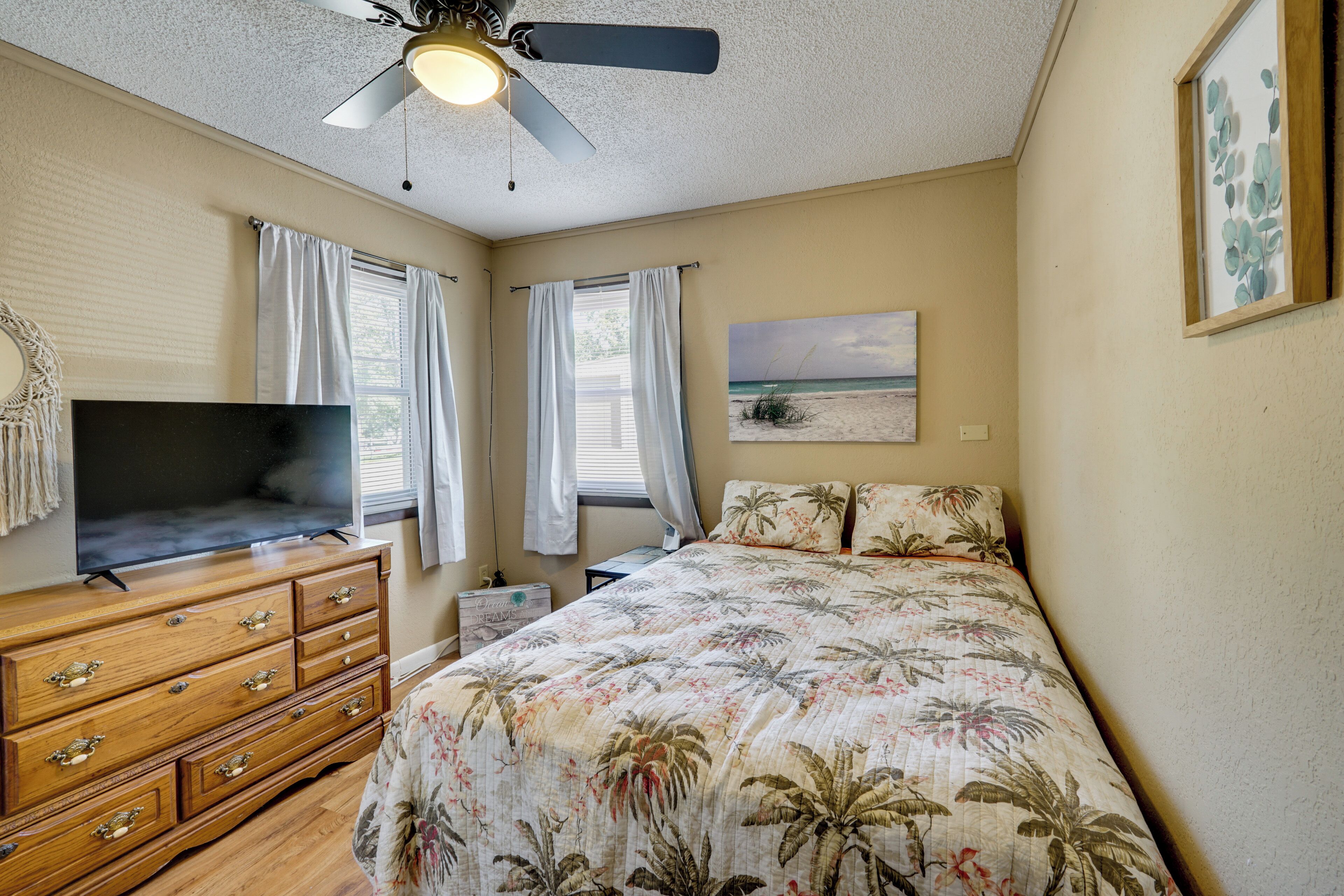 A bedroom with a bed, a TV, a ceiling fan, and a beach-themed painting.