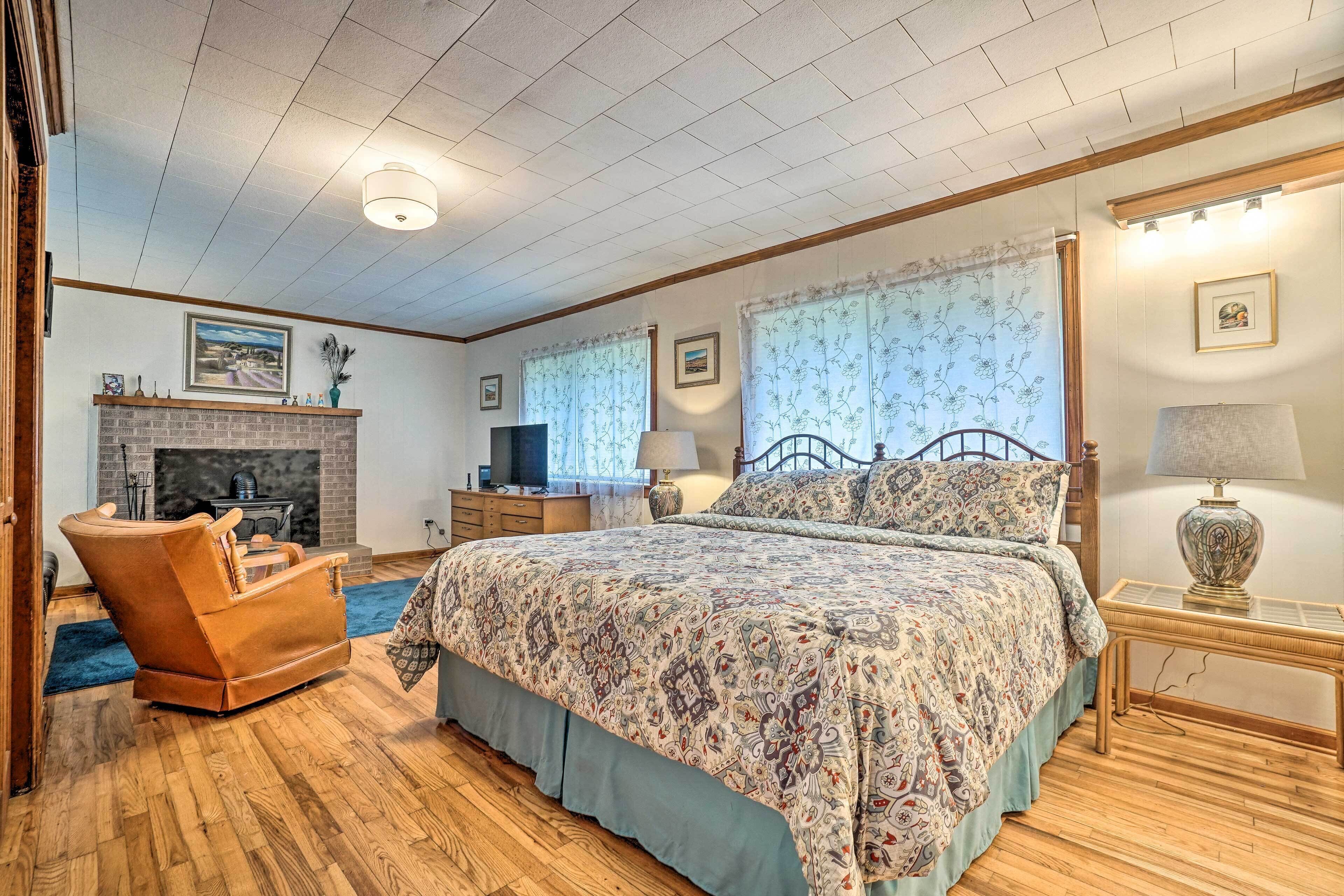 A bedroom with a large bed, a wooden floor, a fireplace, a television, and a window with floral curtains.