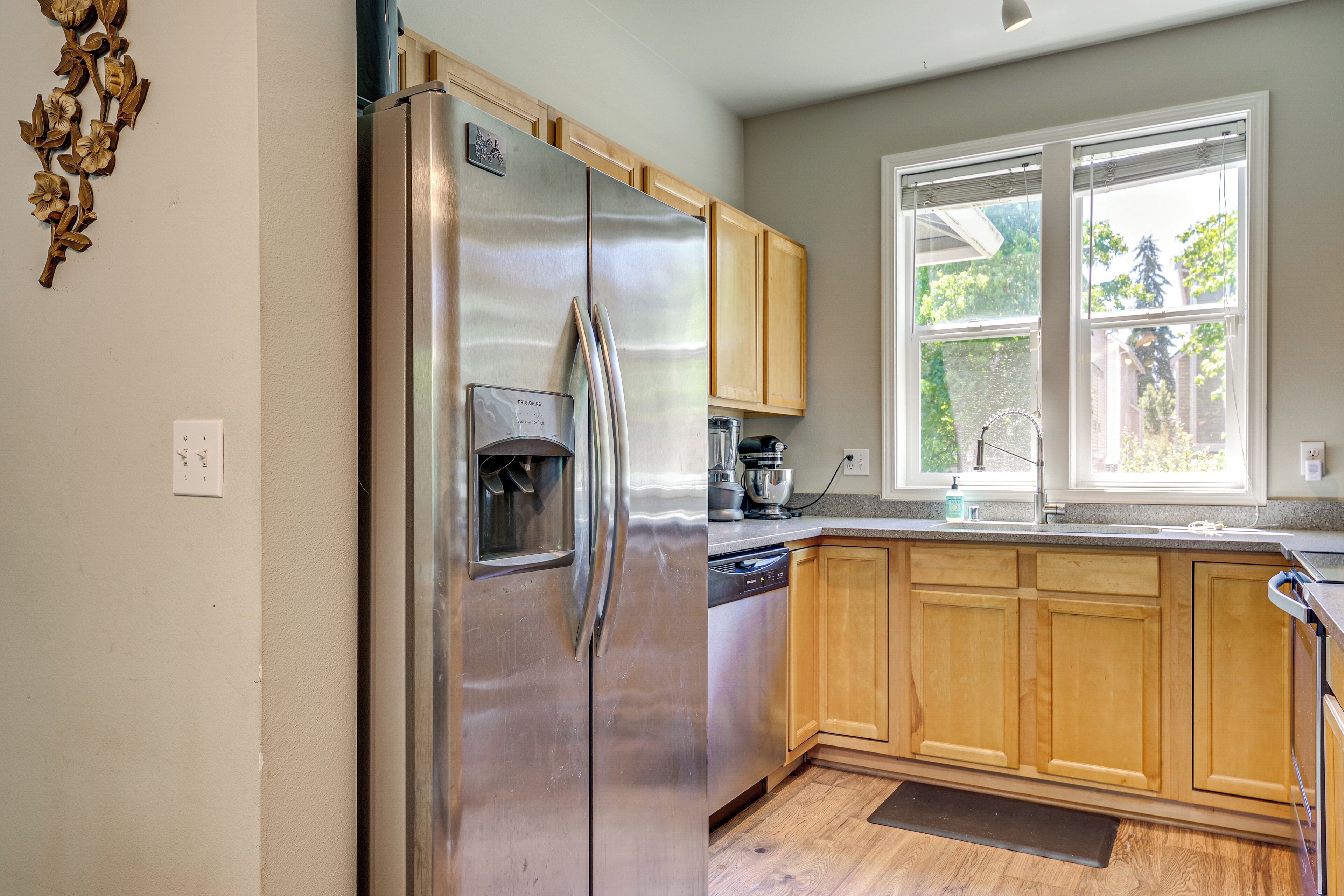 A kitchen with a stainless steel refrigerator, wooden cabinets, a window with blinds, and a dishwasher.