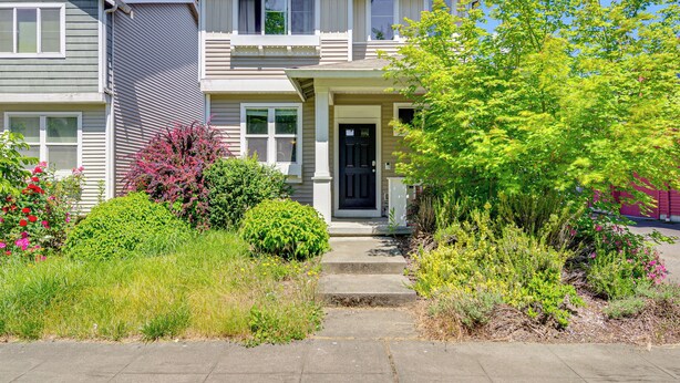 A two-story house with a front porch, a black front door, and a small garden with various plants and flowers. - Half Block to Train! South Beacon Hill Area Home (Seattle)