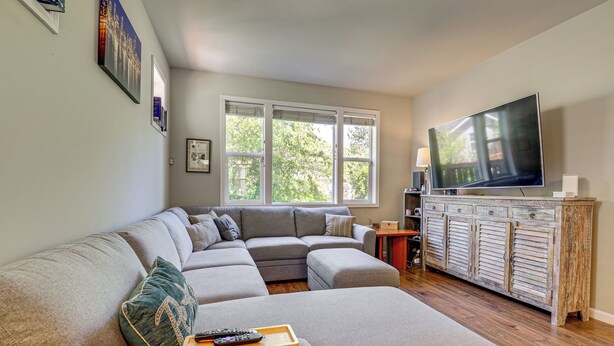 A living room with a sectional sofa, a flat-screen TV, a wooden cabinet, and a window with a view of trees. - Half Block to Train! South Beacon Hill Area Home (Seattle)