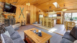 A wooden interior with a TV, fireplace, dining area, kitchen, and a stone wall. - 2 Mi to Big Bear Lake! Family Haven w/ Hot Tub (Big Bear City)
