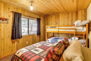 Ein Zimmer in einer Holzhütte mit Etagenbetten, einer gesteppten Bettdecke, einem Fenster mit Vorhängen und einer Pendelleuchte.