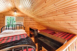 Eine Hütte mit geneigtem Dach, einem Bett mit rot-schwarzer Karodecke, einem kleinen Tisch und einem Fenster mit Blick auf Bäume.