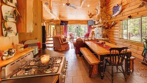 A rustic cabin interior with a wooden ceiling, a dining table, a stove, and a cozy seating area. - Angler Friendly! Pere Marquette Riverfront Retreat (Baldwin)