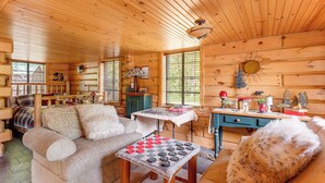 A cozy cabin interior with a wooden ceiling, a sofa, a table with a board game, and a desk with various items. - Angler Friendly! Pere Marquette Riverfront Retreat (Baldwin)