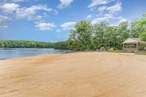 A beach with a sandy area, a wooden structure, and a lake with trees in the background. - Pool & Dock Access! Badin Lake Resort Gem (New London)