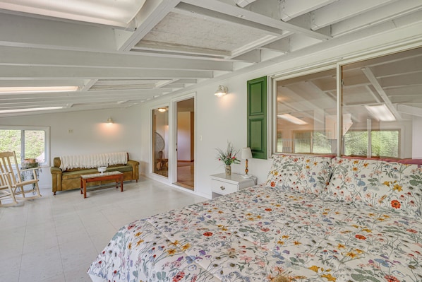 Une pièce spacieuse avec un grand lit, un canapé, un fauteuil à bascule en bois et une petite table avec une lampe. De nombreuses fenêtres laissent entrer la lumière naturelle et offrent une vue sur la verdure environnante.