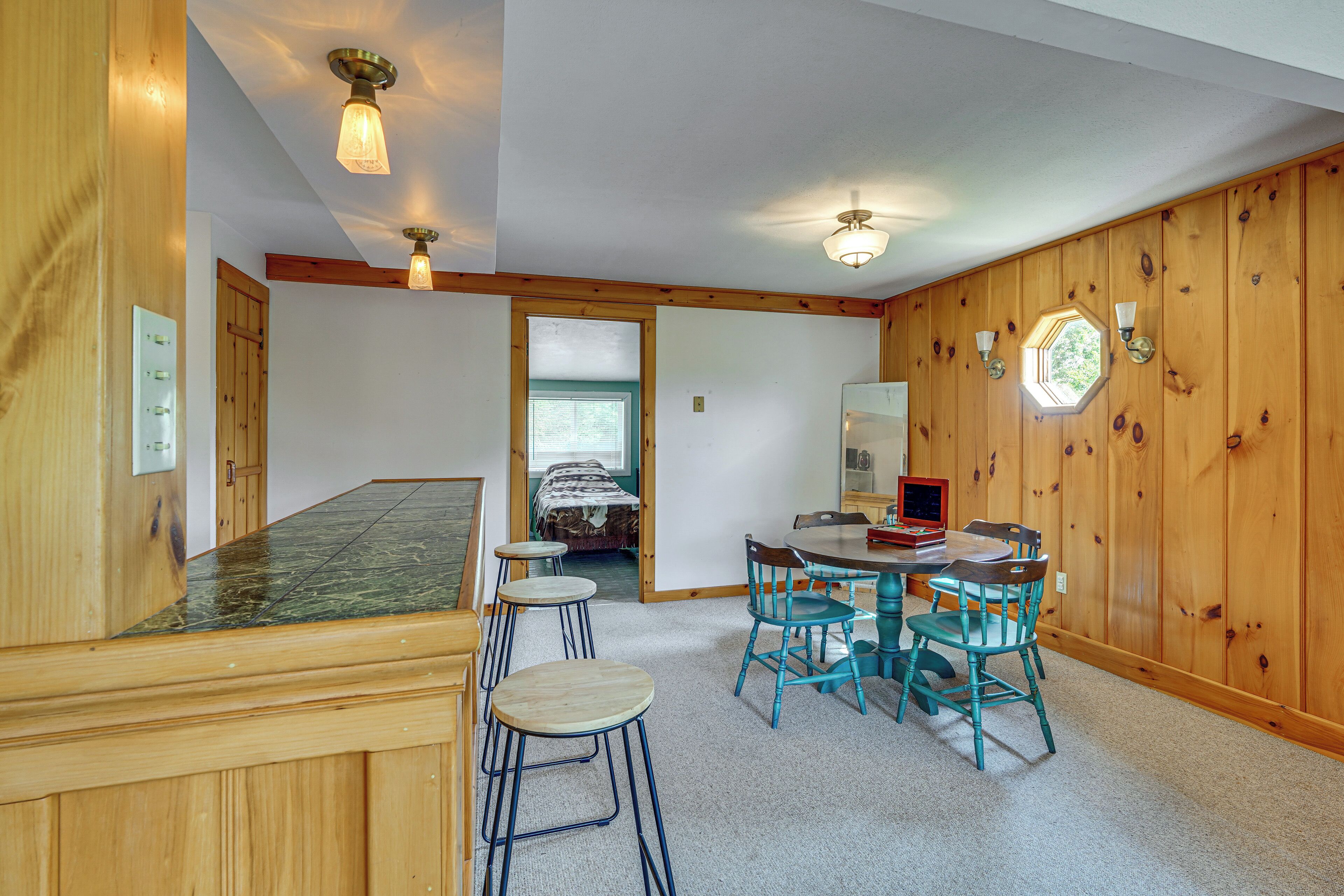 A room with a wooden wall, a dining table with chairs, a kitchen area, and a door leading to another room.