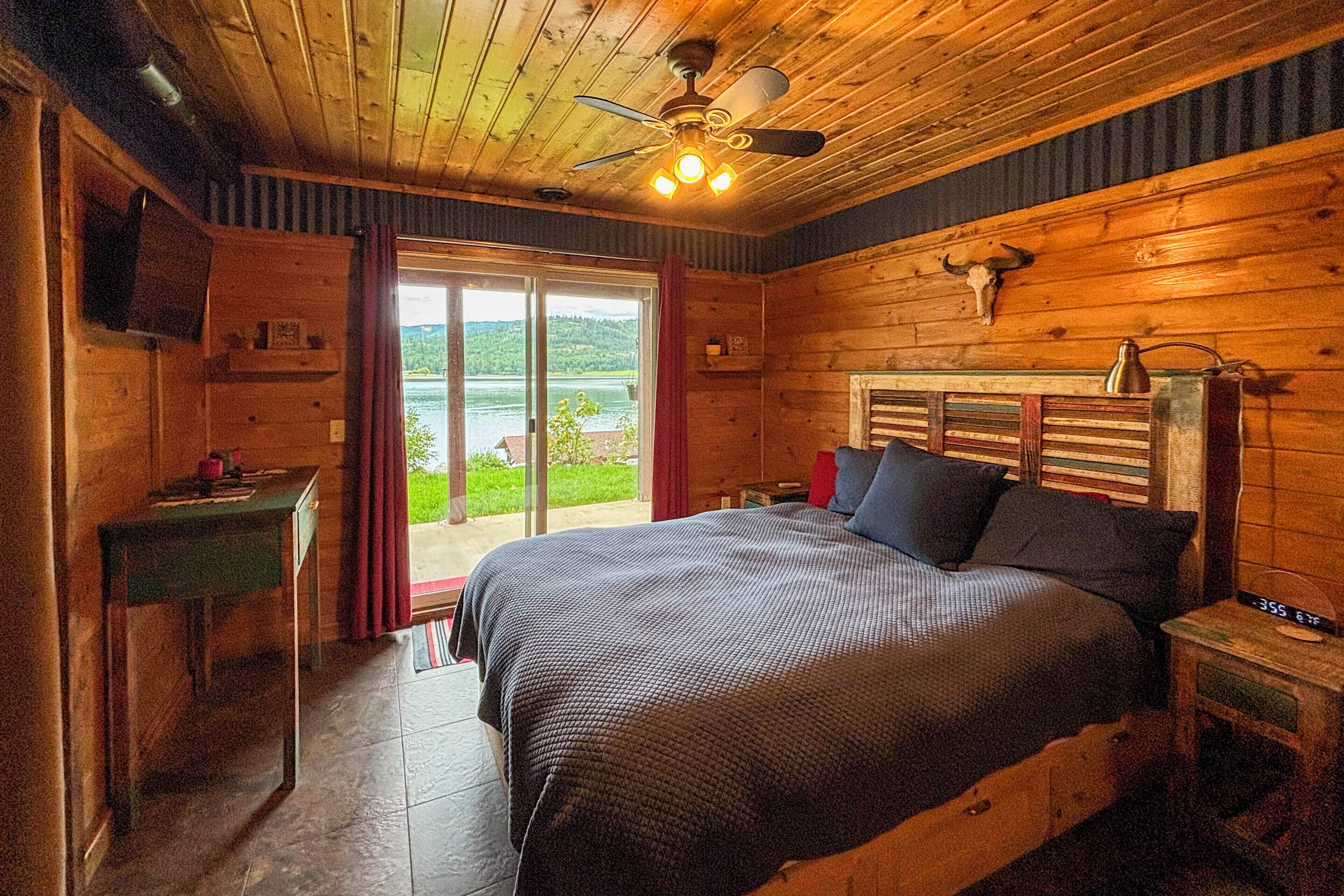 A wooden cabin bedroom with a bed, a desk, a chair, a ceiling fan, and a view of the outdoors.