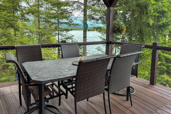 A covered outdoor deck with a table and chairs, overlooking a body of water and surrounded by trees.