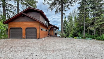 A two-story log cabin with a gravel driveway, surrounded by tall trees.