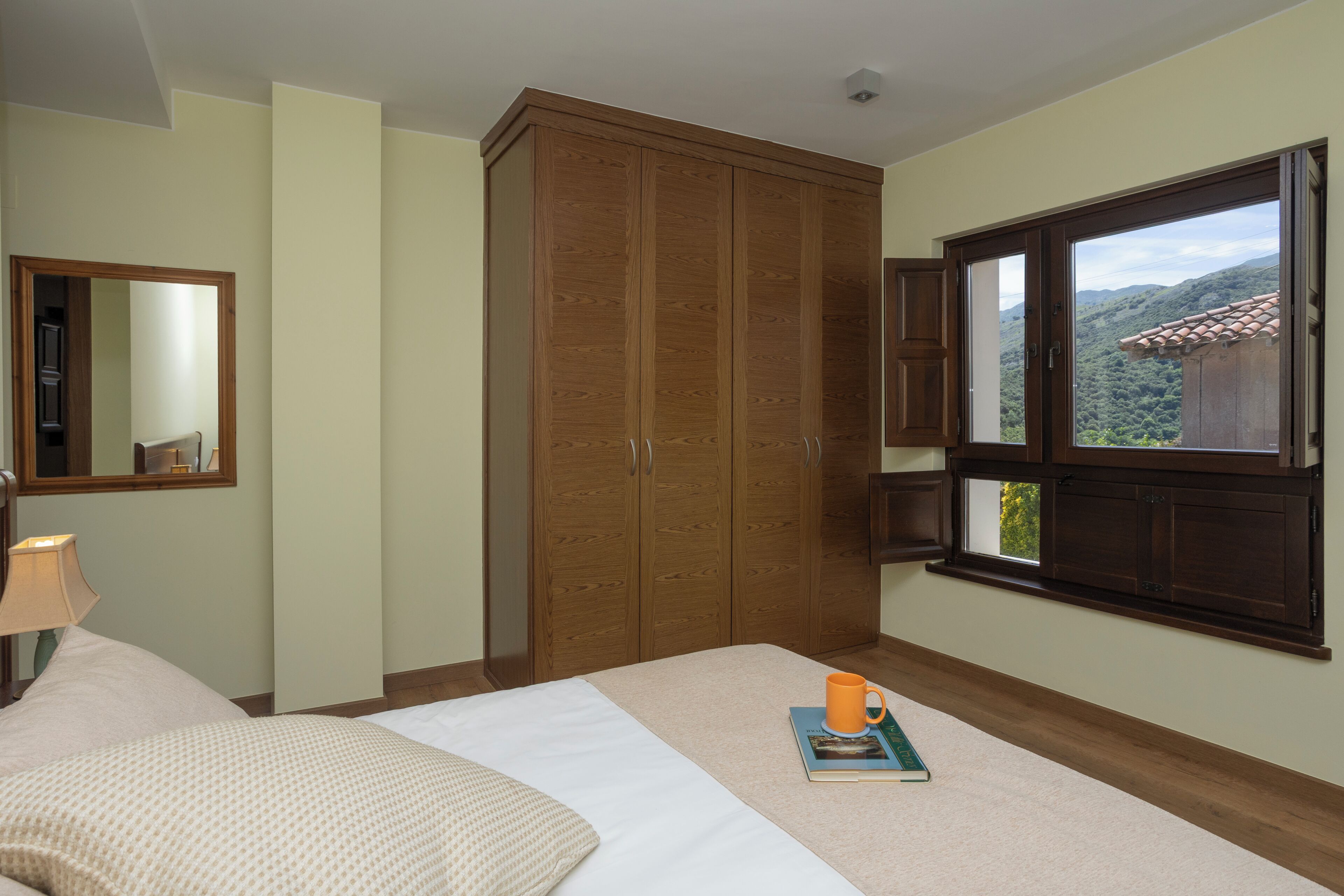 A bedroom with a bed, a wooden wardrobe, a window with a view, and a bedside table with a lamp.