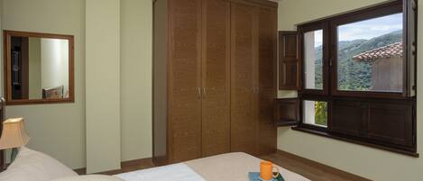 A bedroom with a bed, a wooden wardrobe, a window with a view, and a bedside table with a lamp.