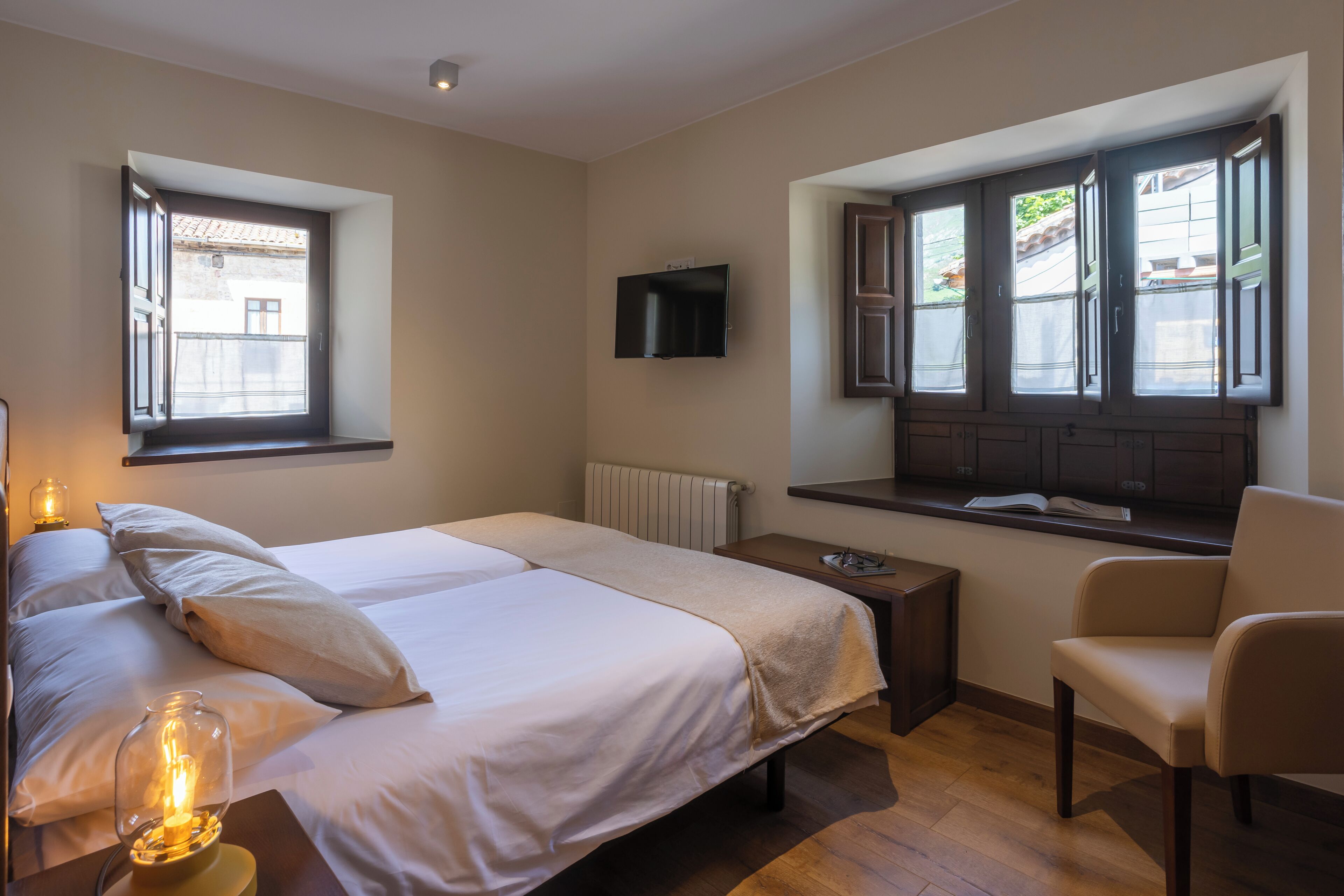 Un dormitorio con dos camas, un televisor, una ventana con contraventanas, una silla y una mesita de noche con lámpara.