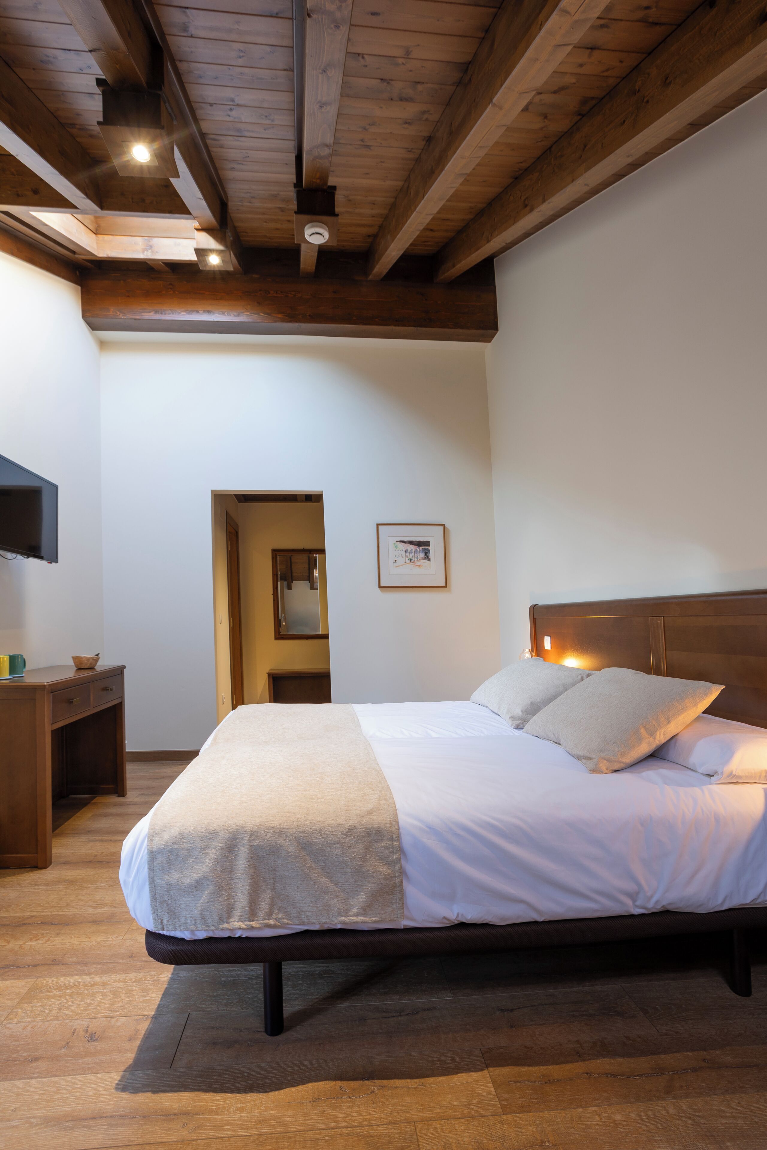 A bedroom with a wooden ceiling, a bed with white bedding, a bedside table, and a wall-mounted TV.