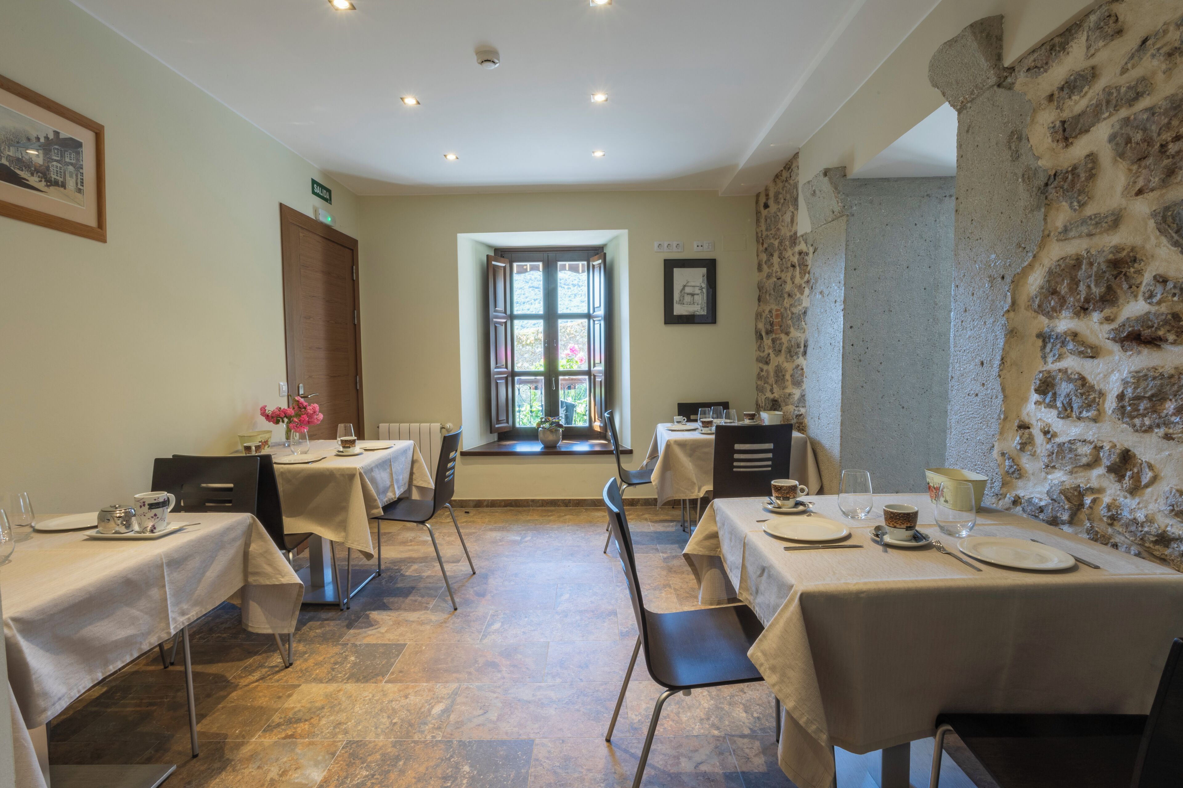 A dining area with tables set for four, a stone wall, and a window with a view.