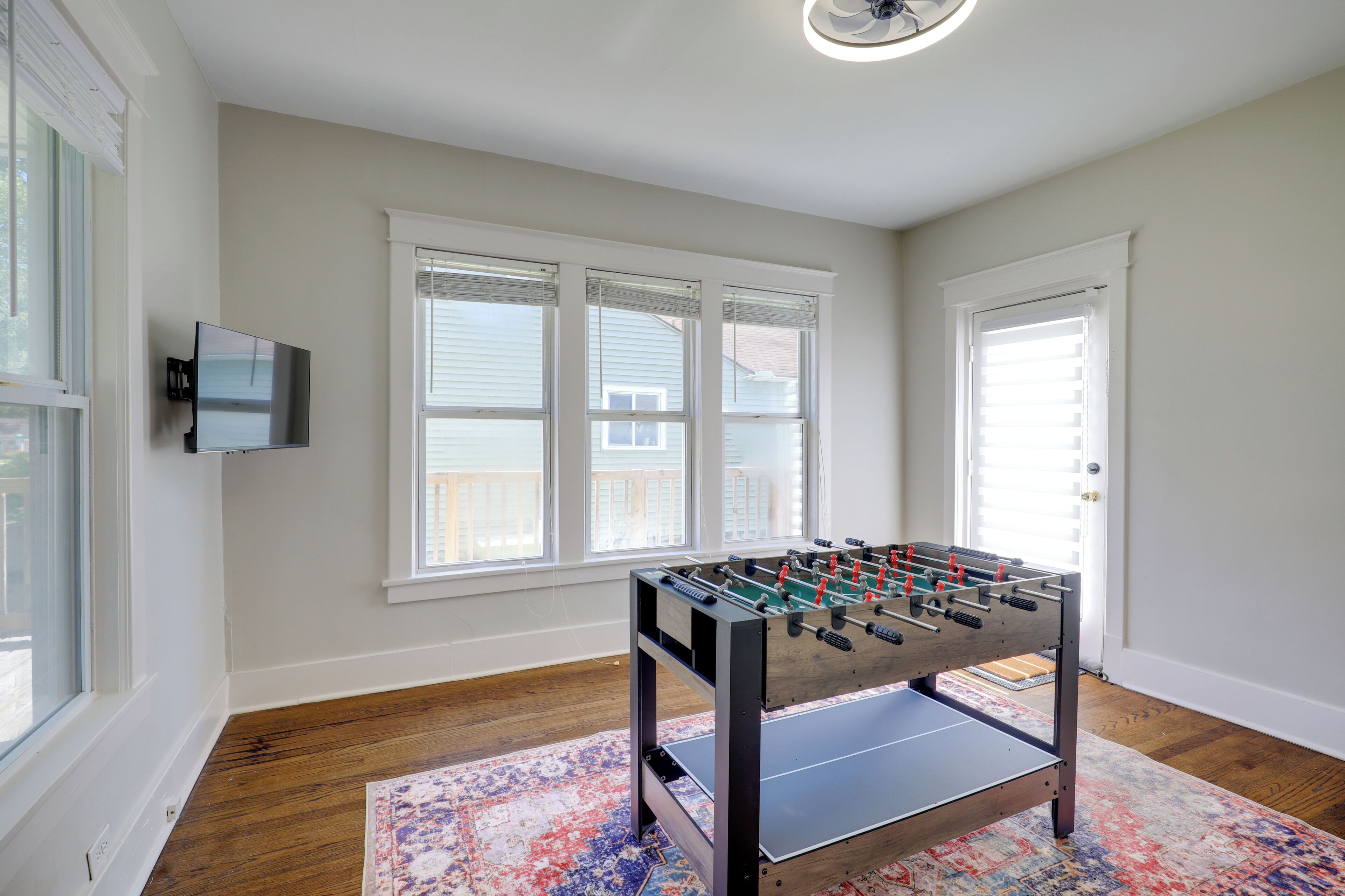 A room with a foosball table, large windows, a television, and a patterned rug.