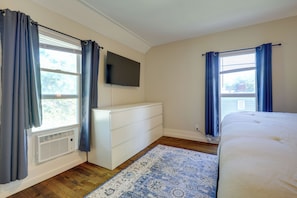 Quarto com cama, penteadeira, televisão e duas janelas com cortinas.