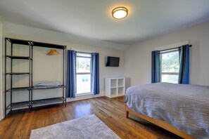 Quarto com cama, piso de madeira, prateleira branca, televisão e janela com cortinas.