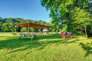 A covered picnic area with a table and chairs, surrounded by a grassy field and trees. - Creek Access & On-site Trails: Serene Ozark Cabin (Stella)