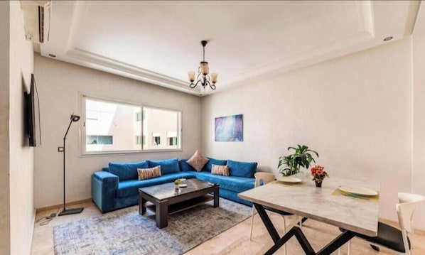 Un salon avec un canapé bleu, une table basse, une table à manger, un lustre et une grande fenêtre.