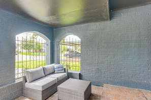Ein überdachter Patio-Bereich mit einem Korbsessel und Hocker, einem Ziegelboden und Blick durch ein vergittertes Fenster.