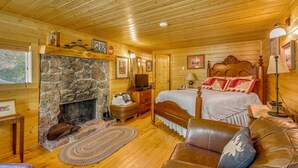 Interieur van een blokhut met een stenen open haard, houten plafond, een bed met hoofdeinde, een leren bank en een televisie.