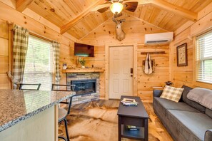 Ein Interieur einer Blockhütte mit Kamin aus Stein, Deckenventilator, montiertem Hirschkopf, einer Couch und einem Essbereich mit Stühlen.