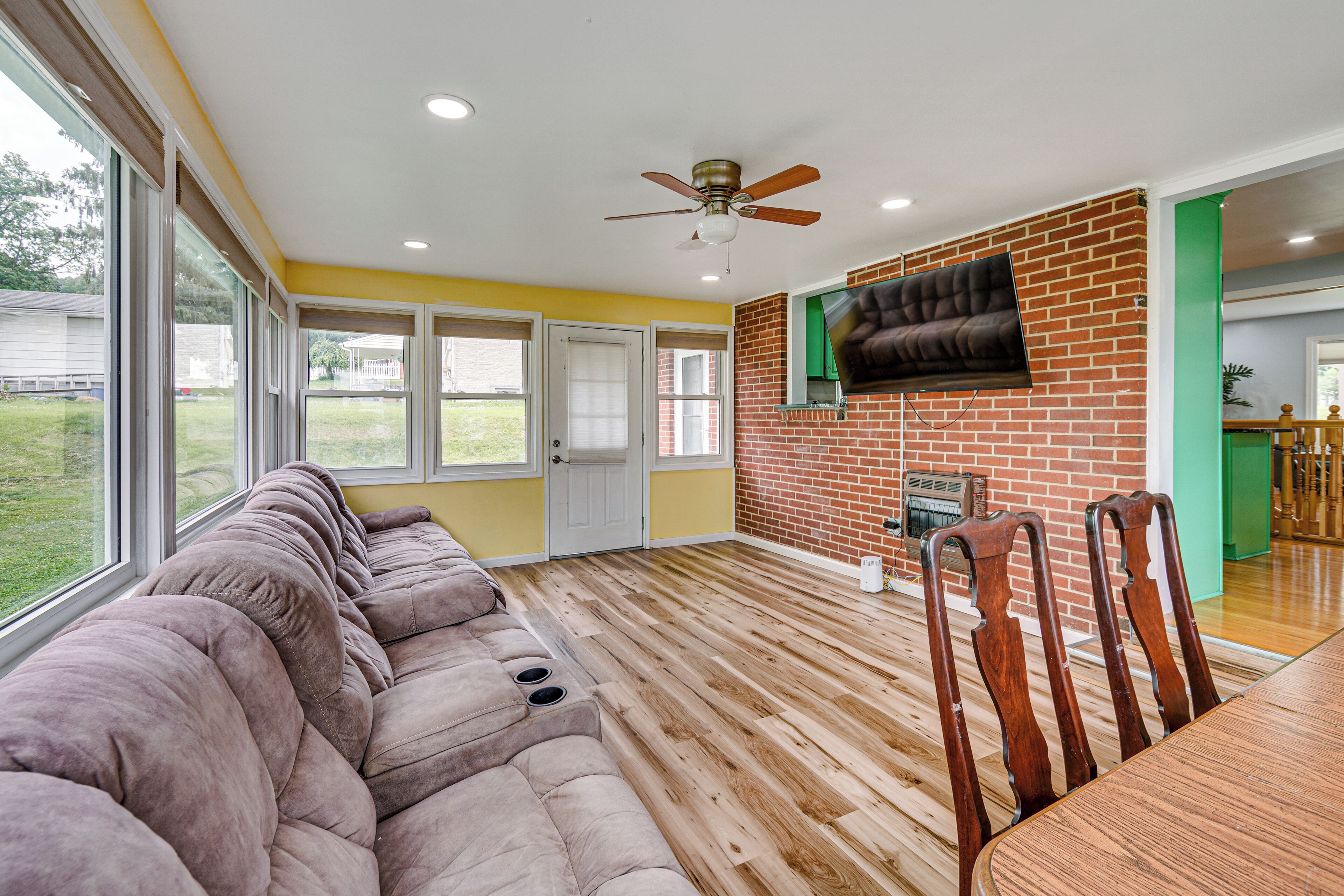 A room with a large sectional sofa, a dining table with chairs, a television mounted on a brick wall, and a ceiling fan.