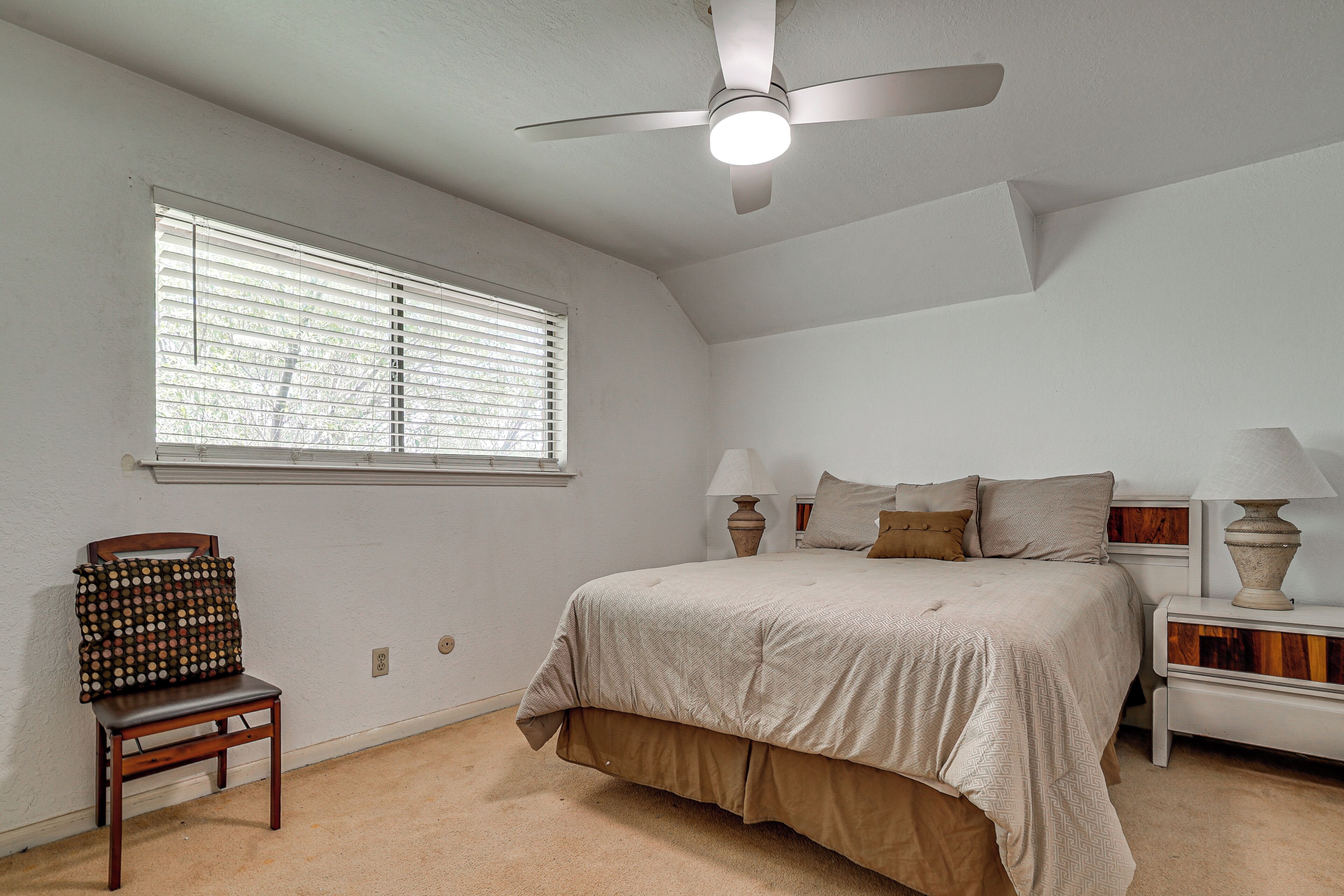 A bedroom with a bed, a chair, a nightstand, a lamp, a ceiling fan, and a window with blinds.