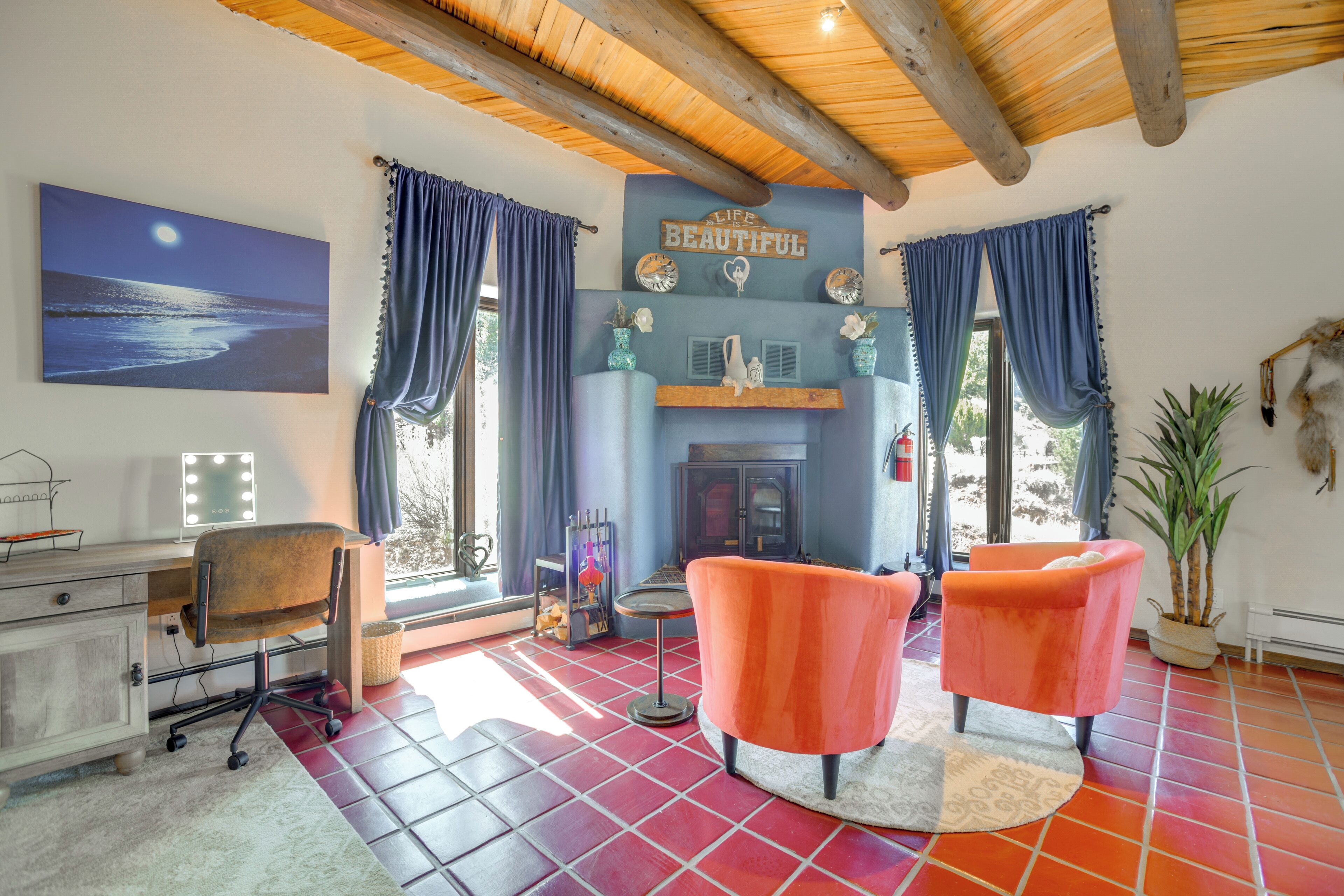 Une pièce avec une cheminée, deux fauteuils orange, un bureau avec un ordinateur et un tableau représentant une scène de plage.