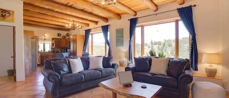 Un salón amplio con dos sofás, una mesa de centro y un ordenador portátil. La habitación tiene techos de madera y ventanas grandes con cortinas.