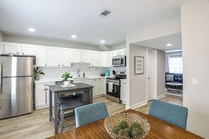 A modern kitchen with white cabinets, stainless steel appliances, and a dining area with a wooden table and blue chairs. - Walk to Stadium and Campus! Cozy Townhome w/ Pool (Oxford)