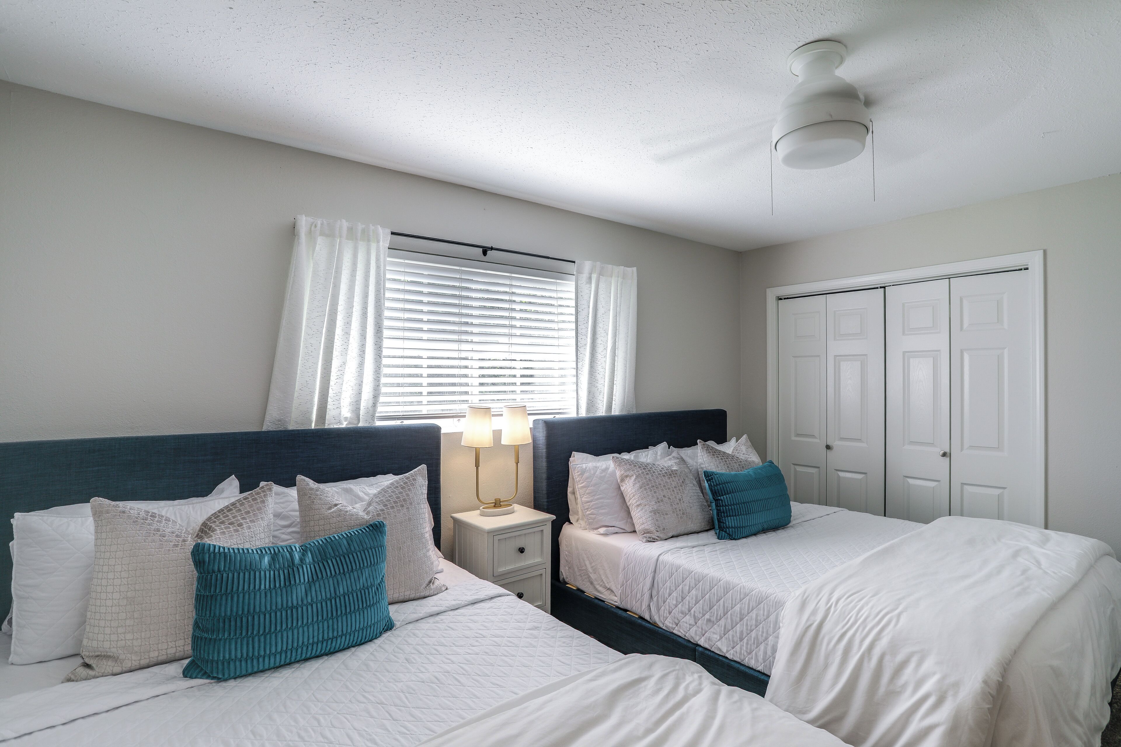 A bedroom with two beds, a nightstand with a lamp, a window with curtains, and a closet.