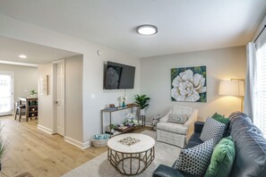 A living room with a sofa, armchair, coffee table, and a TV mounted on the wall. - Walk to Stadium and Campus! Cozy Townhome w/ Pool (Oxford)