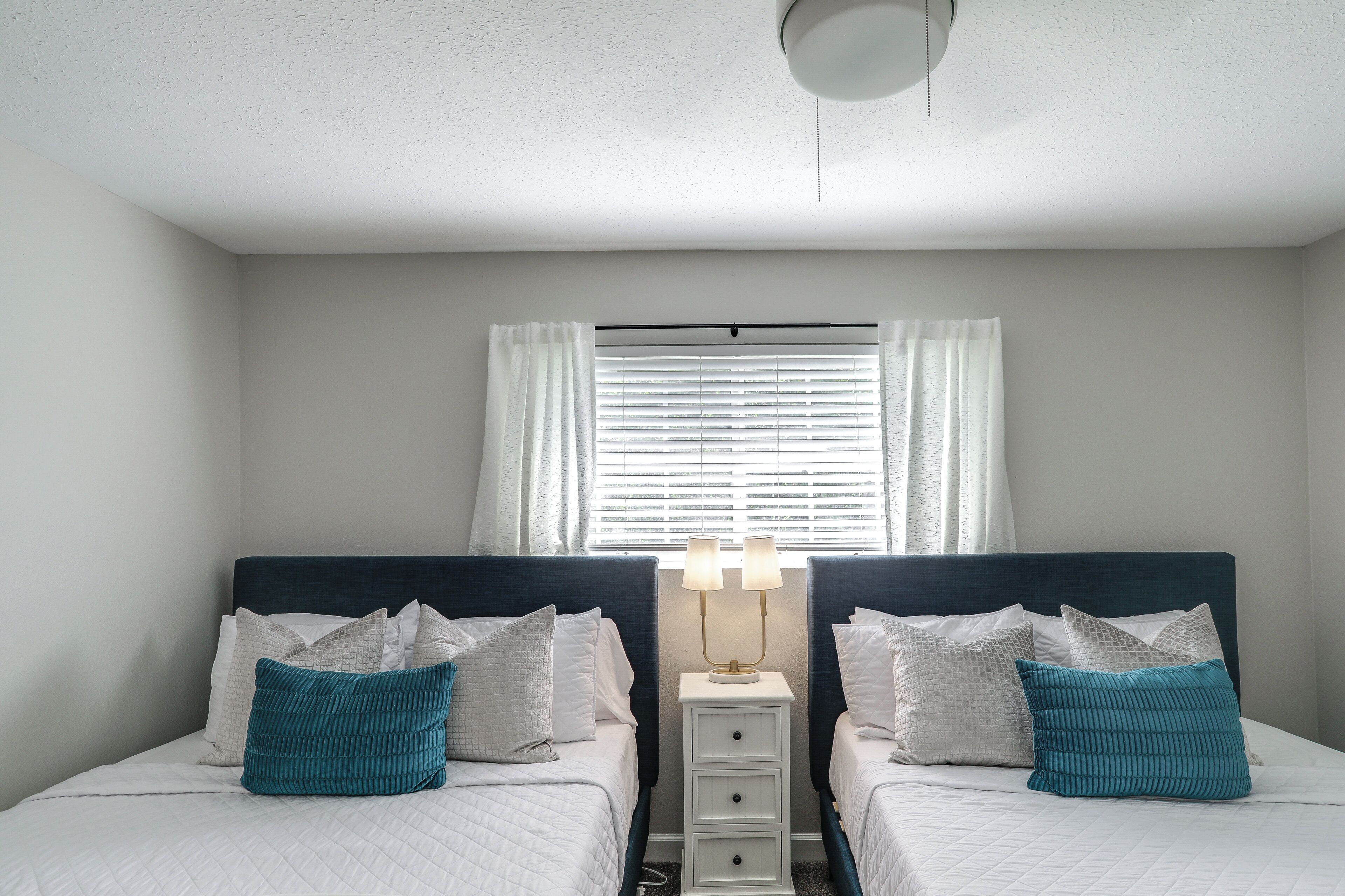 A double bed with white bedding and teal pillows, a bedside table with a lamp, and a window with blinds.