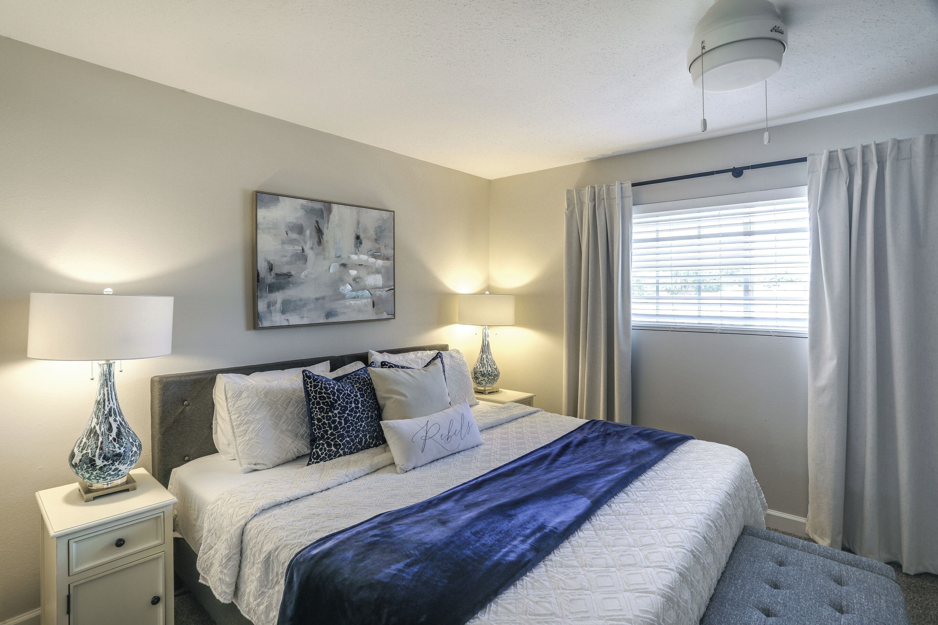 A bedroom with a bed, nightstands, lamps, a painting, and a window with curtains.