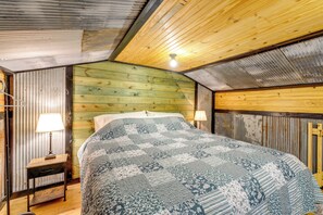 A bedroom with a wooden ceiling, a bed with a patterned bedspread, a nightstand with a lamp, and a wooden wall. - Fish, Hike & Unwind! Rustic Ozark Cabin (Stella)