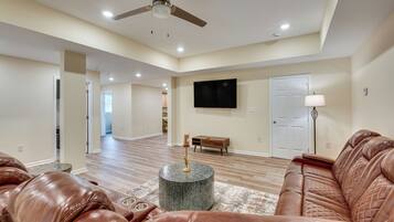 A spacious living room with leather sofas, a round coffee table, a ceiling fan, recessed lighting, a flat-screen TV, and a door with a lamp.